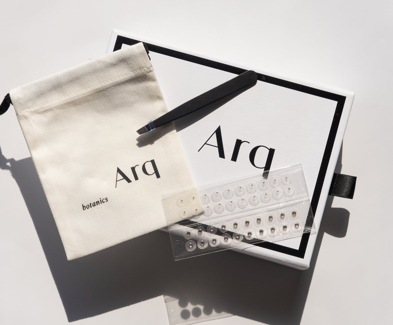 An Arq Botanics Ear Seed Kit, containing a white drawstring pouch, a black pair of tweezers, and multiple sheets of small adhesive seed stickers.