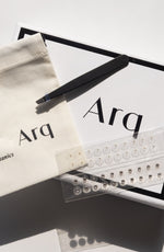 An Arq Botanics Ear Seed Kit, containing a white drawstring pouch, a black pair of tweezers, and multiple sheets of small adhesive seed stickers.