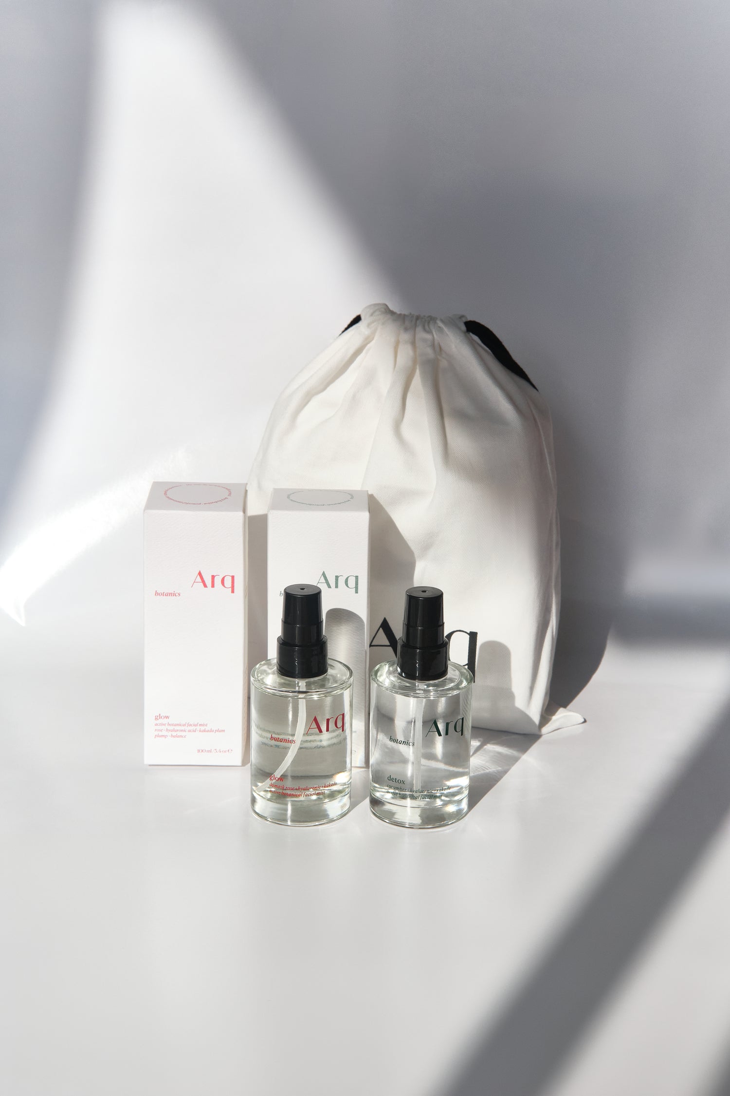 Two clear glass bottles of Arq Botanics Glow Hyaluronic Acid Facial Mist with black spray pumps, next to a white drawstring pouch.