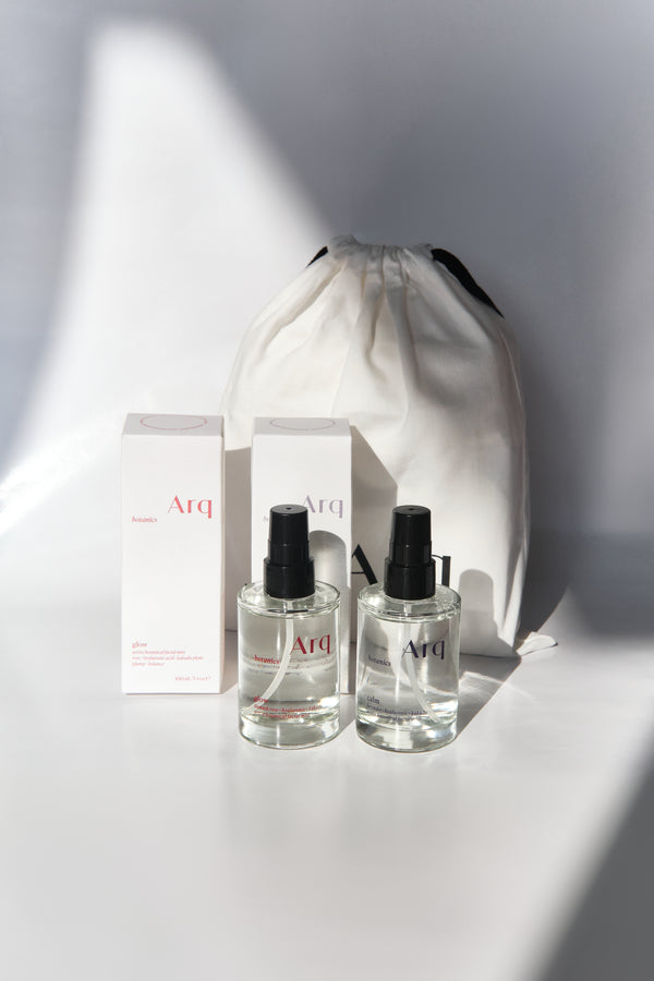 Two clear glass bottles of Arq Botanics Glow Hyaluronic Acid Facial Mist with black spray pumps, next to a white drawstring pouch.