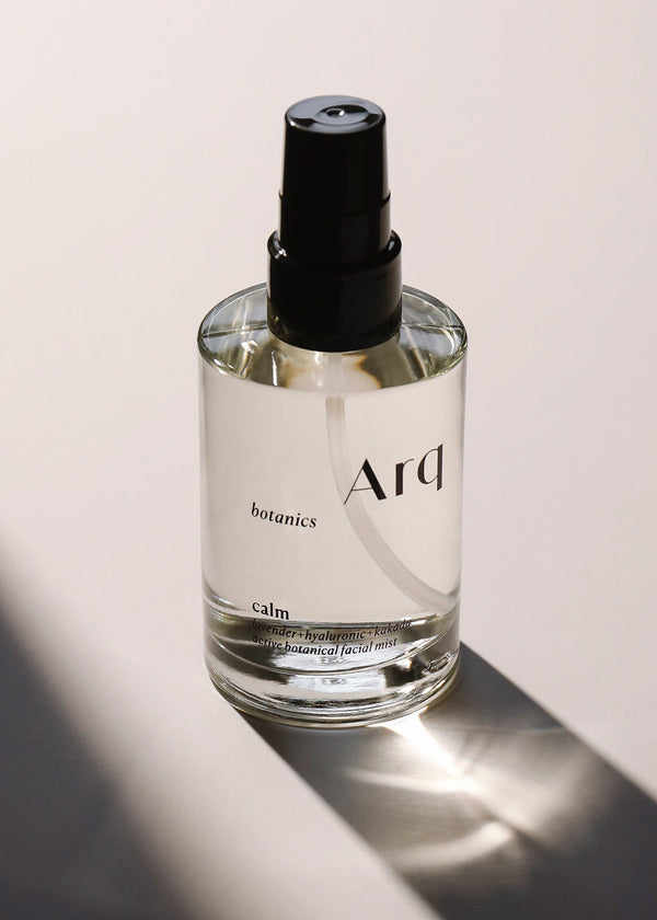 A glass bottle of Arq Botanics Calm Lavender Hyaluronic Kakadu Active Botanical Facial Mist with a black spray pump, sitting on a white surface.