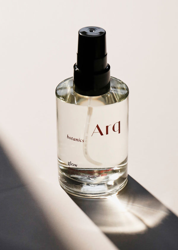 A glass bottle of Arq Botanics Glow Rose Botanical Facial Mist with a black spray pump, sitting on a white surface.